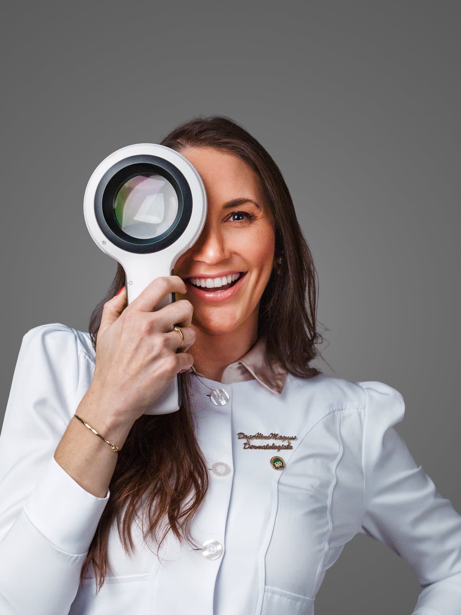 Dra. Aline Magnus sorrindo, com um dermatoscópio diante de um dos olhos