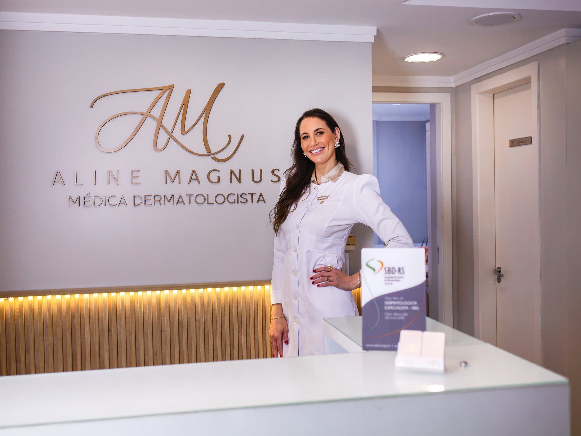 Dra. Aline Magnus na recepção do consultório, diante do logotipo Aline Magnus — Médica Dermatologista