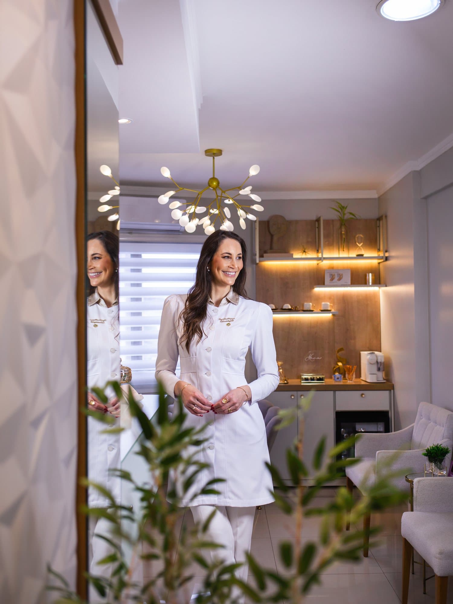 Dra. Aline Magnus em pé na recepção, sob um lustre dourado, com plantas em primeiro plano